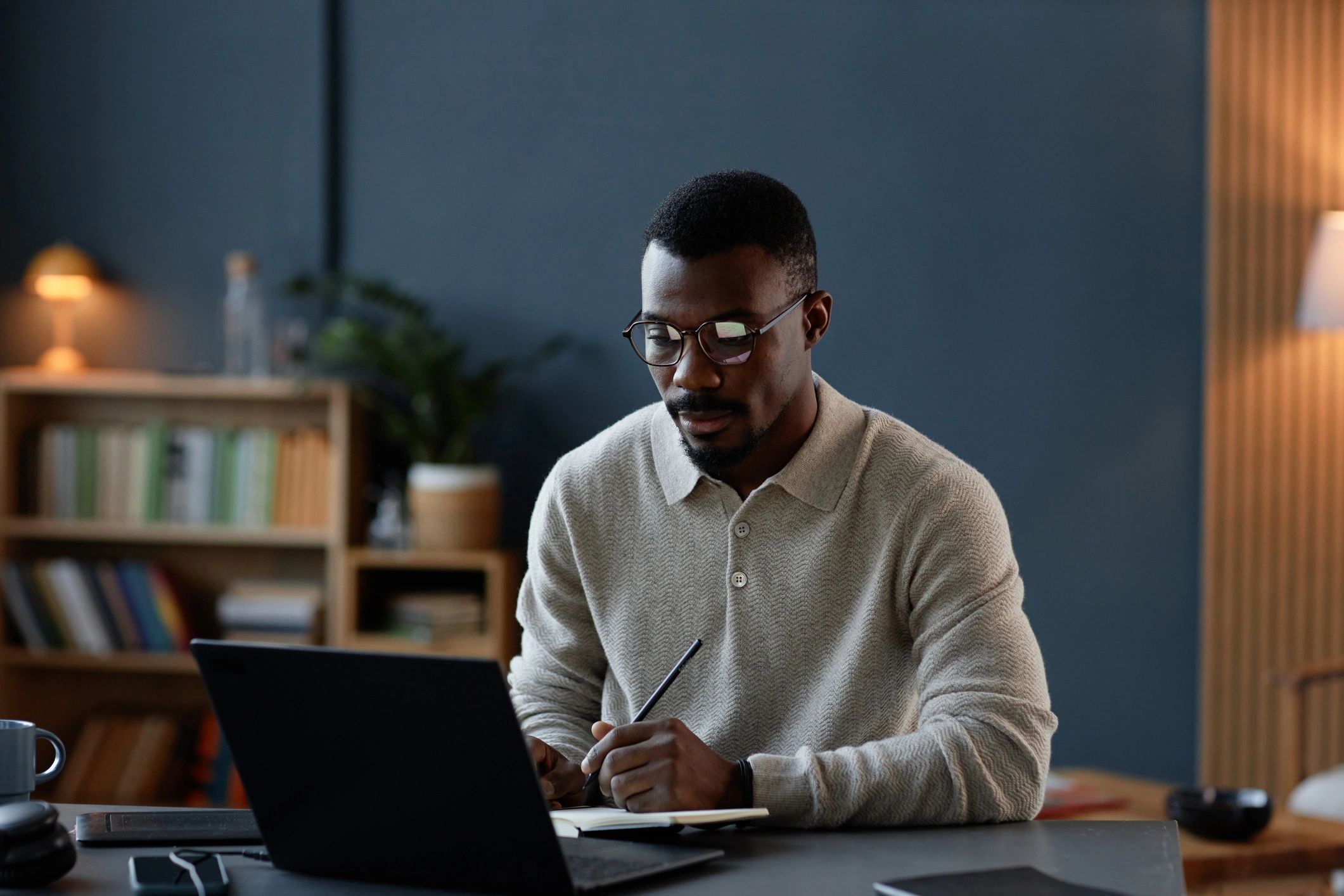 Black Male Interior Designer Writing in Notebook while Using Laptop at Desk in Office
